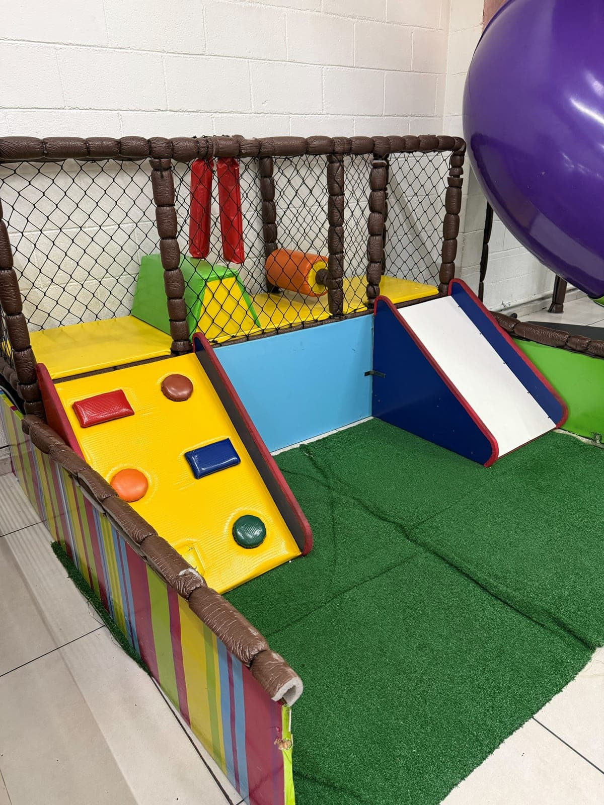 Área baby cercada, com rampas macias, escorregador e piso de grama sintética