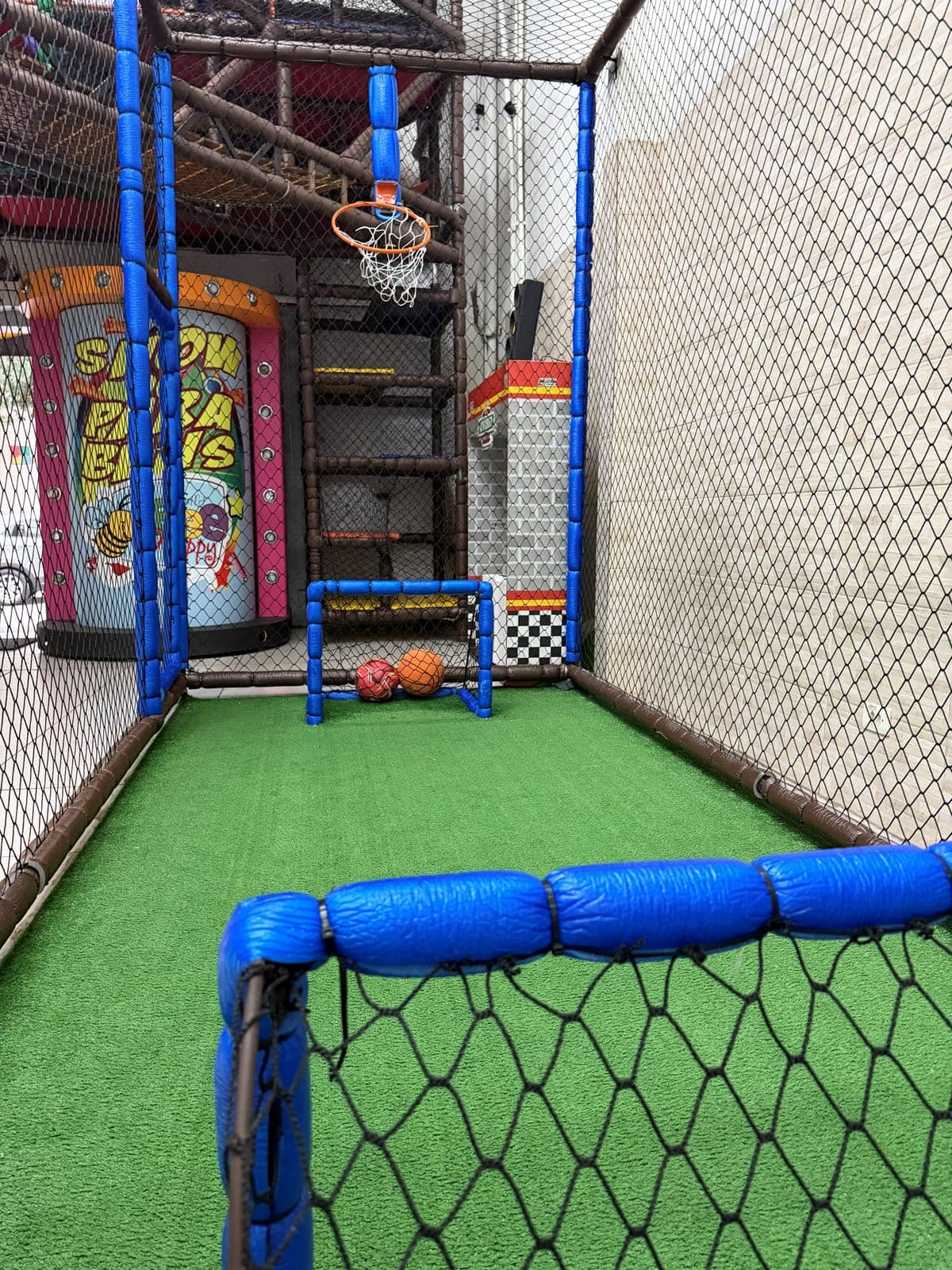 Mini campo de futebol com gol, cesta de basquete e grama sintética, cercado por rede de proteção